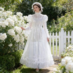 May include: A white, long-sleeved dress with a high collar, puff sleeves, and a layered skirt. The dress features lace detailing and is worn with a matching headpiece. The background includes white roses and a white picket fence.