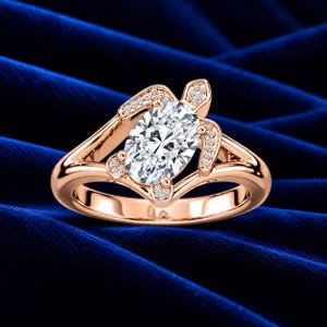 May include: A rose gold ring featuring a central oval-cut white gemstone surrounded by a turtle-shaped setting. The turtle's head, flippers, and shell border are accented with small, clear pavé-set stones along the polished metal band.