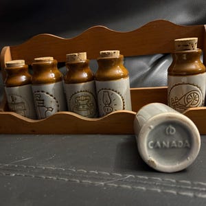 Peut inclure: Un porte-épices vintage avec six pots en céramique. Les pots sont de couleur marron et beige avec des bouchons en liège. Chaque pot est décoré d'un motif en relief différent. Le support en bois est de couleur marron chaud. Le fond d'un pot indique "CANADA".