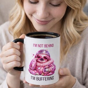 May include: White ceramic mug with a black handle and rim. The mug features a pink sloth wearing heart-shaped sunglasses and a hat, with the text "I'M NOT BEHIND" and "I'M BUFFERING".