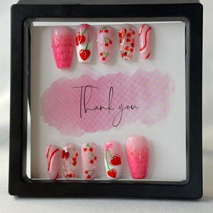 May include: A set of ten artificial nails in a black display case. The nails feature various designs, including cherries, hearts, and pink gradients. The words "Thank you" are written on a pink background.