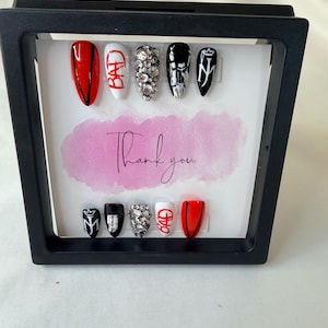 May include: A black framed display of artificial nails. The nails feature various designs, including red, white, and black colors, rhinestones, and the word "BAD". A pink watercolor background with the words "Thank you" is visible.