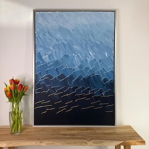 Può includere: Dipinto astratto in cornice argentata. L'opera presenta un design strutturato e stratificato in tonalità di blu, con riflessi bianchi e accenti dorati. Il dipinto è esposto su un tavolo di legno, con un vaso di tulipani a sinistra.