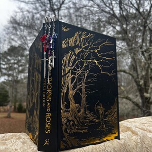 Op de afbeelding: Een zwart boek met een gouden boom- en figuurontwerp, getiteld "Thorns and Roses Collector's Edition". Drie zwaardvormige bladwijzers met kleurrijke kralen hangen aan de bovenkant. Het boek wordt gepresenteerd op een kanten oppervlak.