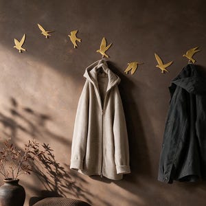 May include: A set of six gold-colored bird-shaped wall hooks mounted in a horizontal arrangement. A beige hooded zip-up jacket hangs from one hook, and a dark gray hooded jacket hangs from another. The hooks are shaped like birds in flight.