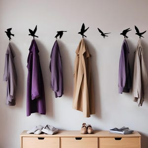 May include: A modern entryway with a light wood cabinet, a purple sofa, and a round mirror. Black bird-shaped hooks hold coats in shades of purple and tan. A stylish and functional space.