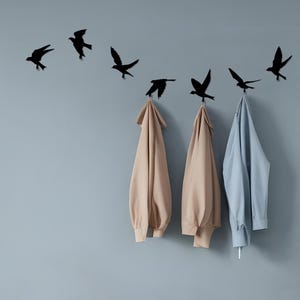 May include: A wall-mounted coat rack featuring black bird silhouettes in flight. Three jackets in beige and light blue hang from the hooks. The background is a solid, light blue-gray color, creating a minimalist aesthetic.