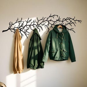 May include: A black metal tree branch wall-mounted coat rack with three jackets hanging from it. The jackets are a tan hoodie, a dark green jacket, and a green hooded jacket. The wall is a light beige color.