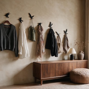 Può includere: Un set di sette ganci da parete neri a forma di uccello montati in fila, ognuno dei quali sostiene vari oggetti tra cui un cardigan grigio scuro, una camicia bianca, un top verde, una sciarpa fantasia, una giacca nera, una borsa beige e un panno a quadri.