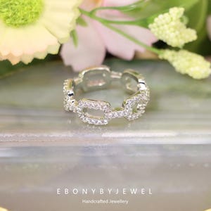 May include: A silver ring featuring a chain link pattern, adorned with tiny, clear gemstones. The ring is displayed against a blurred backdrop with flowers. The text "EBONY BY JEWEL Handcrafted Jewellery" is present.