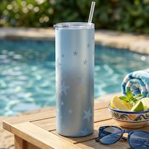 May include: A tall, cylindrical, light blue ombre tumbler with a white star pattern, a clear lid, and a clear straw. The tumbler features a smooth, metallic finish that transitions from a lighter shade at the top to a darker shade at the base.