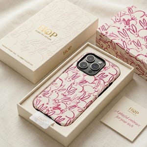 May include: A smartphone case with a magenta rabbit pattern on a cream background, housed in a matching cream box with gold text reading HOP RABBITRY DESIGN. A small card with the text A playful embrace for your tech is included in the box.
