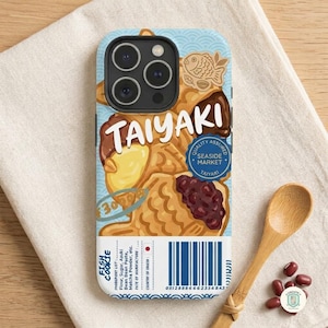May include: A phone case featuring a graphic design of fish-shaped taiyaki pastries with red bean and custard fillings. The design includes the text TAIYAKI, a blue circular seal, a barcode, and a list of ingredients printed on a white background.