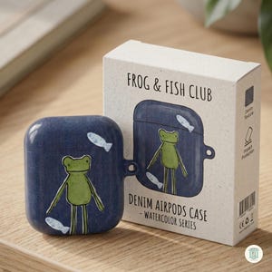 May include: A dark blue AirPods case with a denim texture and a green frog and white fish illustration. The packaging box is labeled FROG & FISH CLUB, DENIM AIRPODS CASE - WATERCOLOR SERIES, and features icons for denim texture and impact protection.