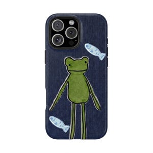 May include: A dark blue denim-textured phone case featuring a hand-drawn style illustration of a green frog standing in the center. Two small, light blue fish are positioned diagonally, one above the frog to the right and one below to the left.