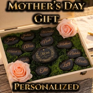 May include: A wooden box with a Mother's Day gift arrangement. The box contains personalized black stones with names, surrounded by green moss and two pink roses. The words "MOTHER'S DAY GIFT" and "PERSONALIZED" are displayed.