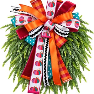 May include: A decorative wreath featuring a large multi-layered bow made of ribbons in orange, red, teal, and white with pumpkin patterns, plaid, polka dots, and black wavy lines, set against a backdrop of lush green fern-like foliage.