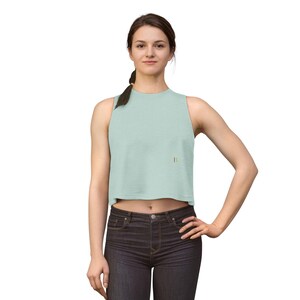 May include: A light green crop top with a gold letter "B" on the front. The sleeveless top is paired with dark wash jeans. The model is standing with one hand on their hip.