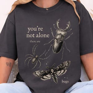 Pode incluir: Camiseta cinza escuro com um gráfico de aranha, besouro e mariposa. O texto "you're not alone there are bugs" é impresso acima e abaixo das ilustrações de insetos. A camiseta é feita de um material macio.