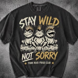 May include: Dark gray t-shirt featuring three cartoon frogs with mohawks sitting on a log. The text "STAY WILD NOT SORRY" is above and below the frogs, with "PUNK ROCK FROGS CLUB" below. The shirt has a vintage, distressed look.