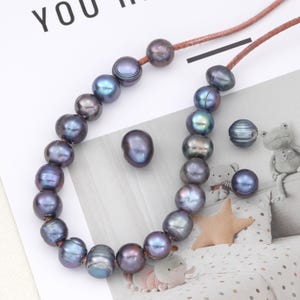 Könnte beinhalten: Halskette mit irisierenden dunkelgrauen Perlen auf einer braunen Kordel. Lose Perlen und eine dekorative Perle sind ebenfalls zu sehen. Die Perlen haben einen Glanz, der Blau- und Grüntöne reflektiert.