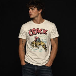 Op de afbeelding: Een gebroken wit t-shirt met korte mouwen en een grafische print van een persoon op een bokkend paard. De tekst CRACK that LASH staat in rood en groen boven de illustratie, die een cowboy in een rood overhemd en een blauwe spijkerbroek toont.