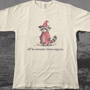 May include: Off-white t-shirt with a raccoon illustration wearing a red cloak and witch's hat. The text "off to commit shenanigans" is printed below the image. A fun, lighthearted design.