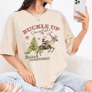 以下が含まれることがあります： クリーム色のTシャツ。テキスト「BUCKLE UP Snowflake」と、トナカイに乗ったカウボーイ、クリスマスツリー、そして「HOWDY CHRISTMAS」の文字がプリントされています。カジュアルで、お祭り気分を盛り上げるデザインです。