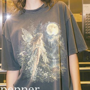 Può includere: Una t-shirt grigio antracite a maniche corte con una stampa grafica dettagliata di una fata seduta in una radura boscosa sotto una luna piena. L'illustrazione include una cascata, un cervo ed elementi floreali. La parola pepper è stampata in bianco.