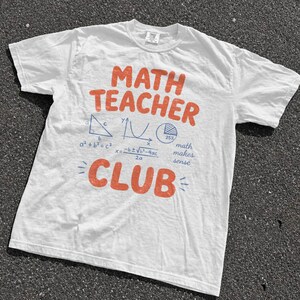 May include: A white short-sleeve t-shirt featuring the orange text MATH TEACHER CLUB. Below the text are blue mathematical graphics including a right triangle, a parabola, a pie chart, the Pythagorean theorem, the quadratic formula, and the text math makes sense.