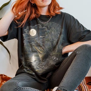 May include: A black short-sleeve t-shirt featuring a graphic print of an alligator with its mouth open in a swampy environment under a full moon. The scene includes dark trees and water reflections, creating a detailed nocturnal landscape illustration.
