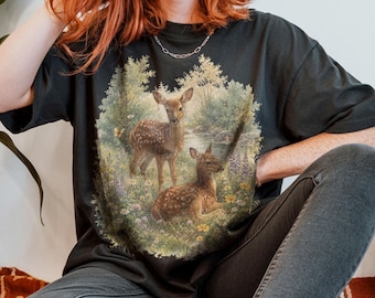 May include: A black short-sleeved t-shirt featuring a detailed graphic print of two spotted fawns in a lush forest setting with wildflowers, greenery, and a small stream. The artwork is centred on the chest, showcasing a natural woodland scene.
