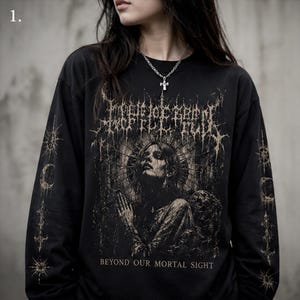 May include: A black long-sleeve shirt featuring a beige graphic print of a person with hands clasped in prayer, surrounded by celestial symbols and the text "BEYOND OUR MORTAL SIGHT." The sleeves are decorated with vertical rows of star and moon icons.