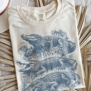May include: A cream-coloured short-sleeve t-shirt featuring a blue graphic print of three alligators in a swampy habitat. The shirt has a crew neckline and a Comfort Colors brand label printed on the inside of the collar area at the neck.