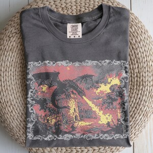 May include: A charcoal gray t-shirt featuring a graphic print of a black dragon perched on a stone tower, breathing yellow fire toward figures on a castle wall. The design is enclosed in an ornate white frame. The neck label reads Comfort Colors.