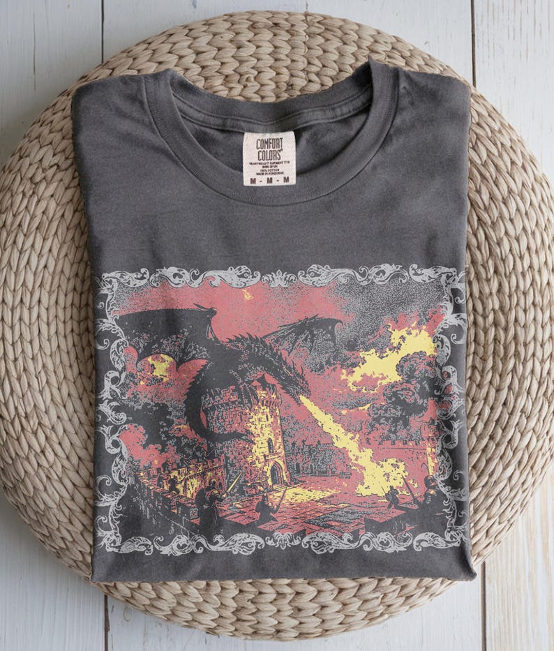 May include: A charcoal gray t-shirt featuring a graphic print of a black dragon perched on a stone tower, breathing yellow fire toward figures on a castle wall. The design is enclosed in an ornate white frame. The neck label reads Comfort Colors.