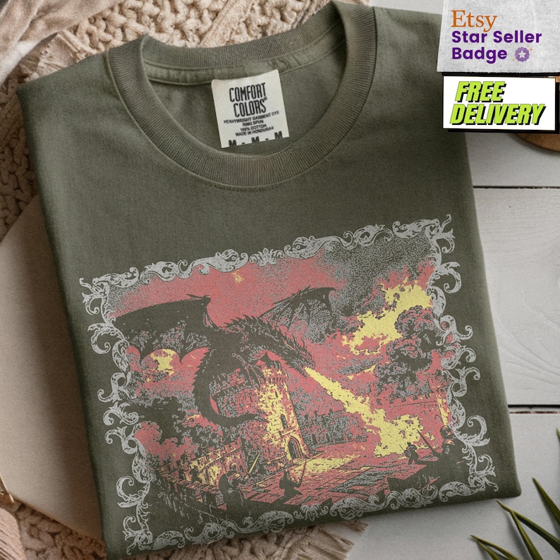 May include: An olive green crew neck t-shirt featuring a graphic print of a black dragon breathing yellow fire at a stone tower. The illustration is enclosed in a decorative white filigree border. The inner neck label reads Comfort Colors.