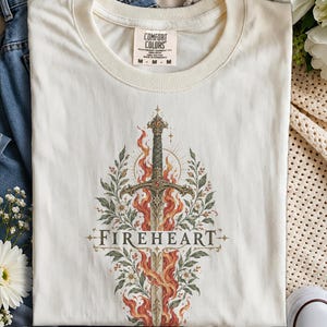 May include: An ivory crew neck t-shirt featuring a graphic print of a sword surrounded by orange flames and green foliage. The word FIREHEART is printed in black capital letters across the centre of the sword. The shirt has a Comfort Colors label.