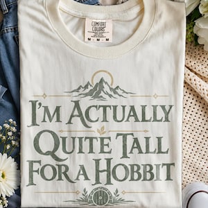 Könnte beinhalten: Ein elfenbeinfarbenes T-Shirt mit Rundhalsausschnitt und einem grünen Grafikdruck, der ein Gebirge, eine runde Tür und den Text "I'M ACTUALLY QUITE TALL FOR A HOBBIT" zeigt. Das Comfort Colors Etikett am Halsausschnitt weist die Größe Medium aus.
