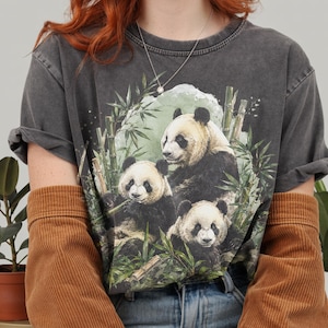 Puede incluir: Una camiseta de manga corta gris carbón con un estampado gráfico de tres pandas gigantes sentados entre tallos y hojas de bambú verdes. La camiseta tiene un cuello redondo y un aspecto de tela ligeramente desgastado.