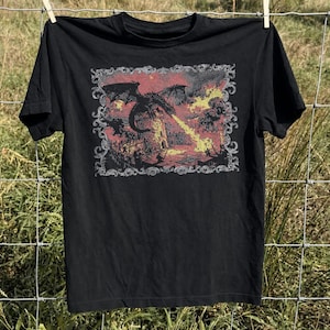 May include: A black short-sleeve t-shirt featuring a rectangular graphic print of a dragon breathing fire onto a stone tower. The illustration is enclosed in an ornate, decorative border and uses a color palette of muted red, yellow, and black tones.