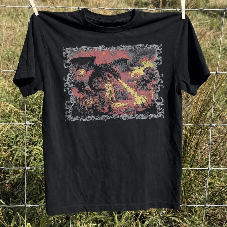 May include: A black short-sleeve t-shirt featuring a rectangular graphic print of a dragon breathing fire onto a stone tower. The illustration is enclosed in an ornate, decorative border and uses a color palette of muted red, yellow, and black tones.