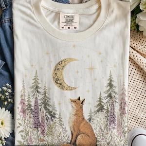 Puede incluir: Una camiseta de color crema con un estampado gráfico de un zorro sentado en un bosque de pinos y flores silvestres moradas. Sobre el zorro hay una gran luna creciente rodeada de pequeñas estrellas. La camiseta tiene una etiqueta Comfort Colors.