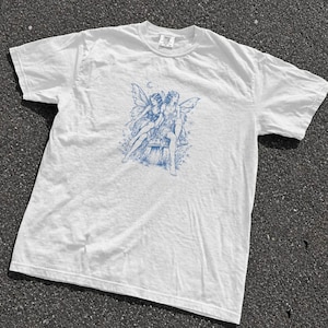 May include: A white short-sleeved t-shirt featuring a blue ink-style graphic print on the chest. The illustration depicts two winged fairy figures sitting together on a large mushroom, surrounded by small stars and a crescent moon above them.