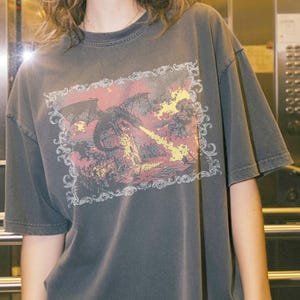 May include: A charcoal gray short-sleeve t-shirt featuring a rectangular graphic print of a black dragon breathing yellow fire onto a stone tower. The illustration is enclosed within an ornate, white, swirling filigree border design.