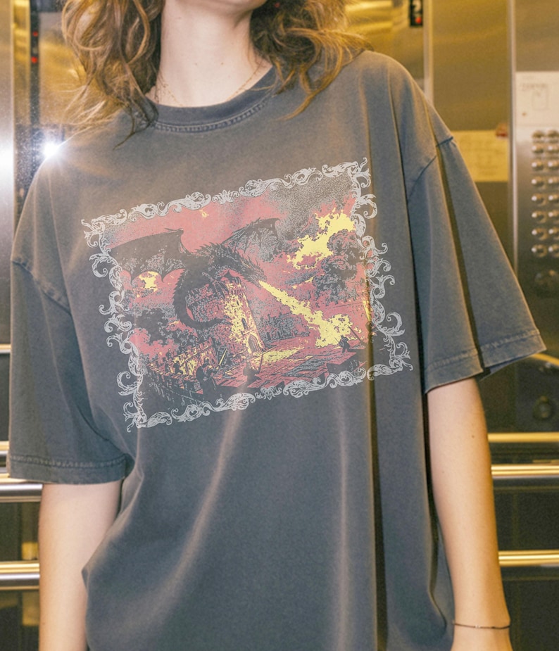 May include: A charcoal gray short-sleeve t-shirt featuring a rectangular graphic print of a black dragon breathing yellow fire onto a stone tower. The illustration is enclosed within an ornate, white, swirling filigree border design.