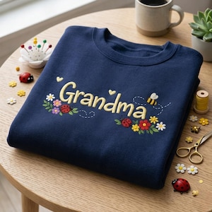 May include: Navy blue sweatshirt embroidered with the word "Grandma" in yellow script, adorned with floral and insect motifs. The design includes ladybugs, flowers, and a bee, creating a charming and personalized gift.