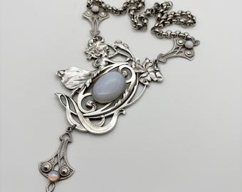 May include: An antique silver necklace featuring an oval, pale blue gemstone pendant. The necklace has intricate floral and Art Nouveau-inspired designs, with a delicate chain and small, round accents. A smaller pendant with a round, opalescent stone hangs below.