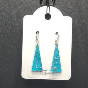 May include: A pair of turquoise resin earrings with silver leaf accents, hanging from a white tag. The earrings are triangular in shape and have a silver hook. The tag has the text "Sure & Luster".