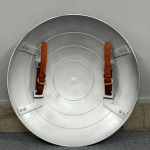 Puede incluir: Un escudo circular de metal plateado con un dise&ntilde;o liso de anillos conc&eacute;ntricos. El interior incluye dos correas de cuero marr&oacute;n con hebillas de metal dorado, cada una asegurada al escudo mediante soportes y remaches de metal plateado.